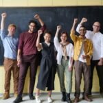 A group of eight adults cheerfully standing with raised fists in front of a blackboard indoors.